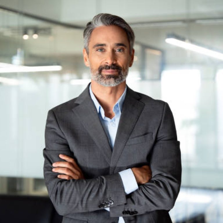 Handsome hispanic senior business man with crossed arms smiling at camera. Indian or latin confident mature good looking middle age leader male businessman on blur office background with copy space.